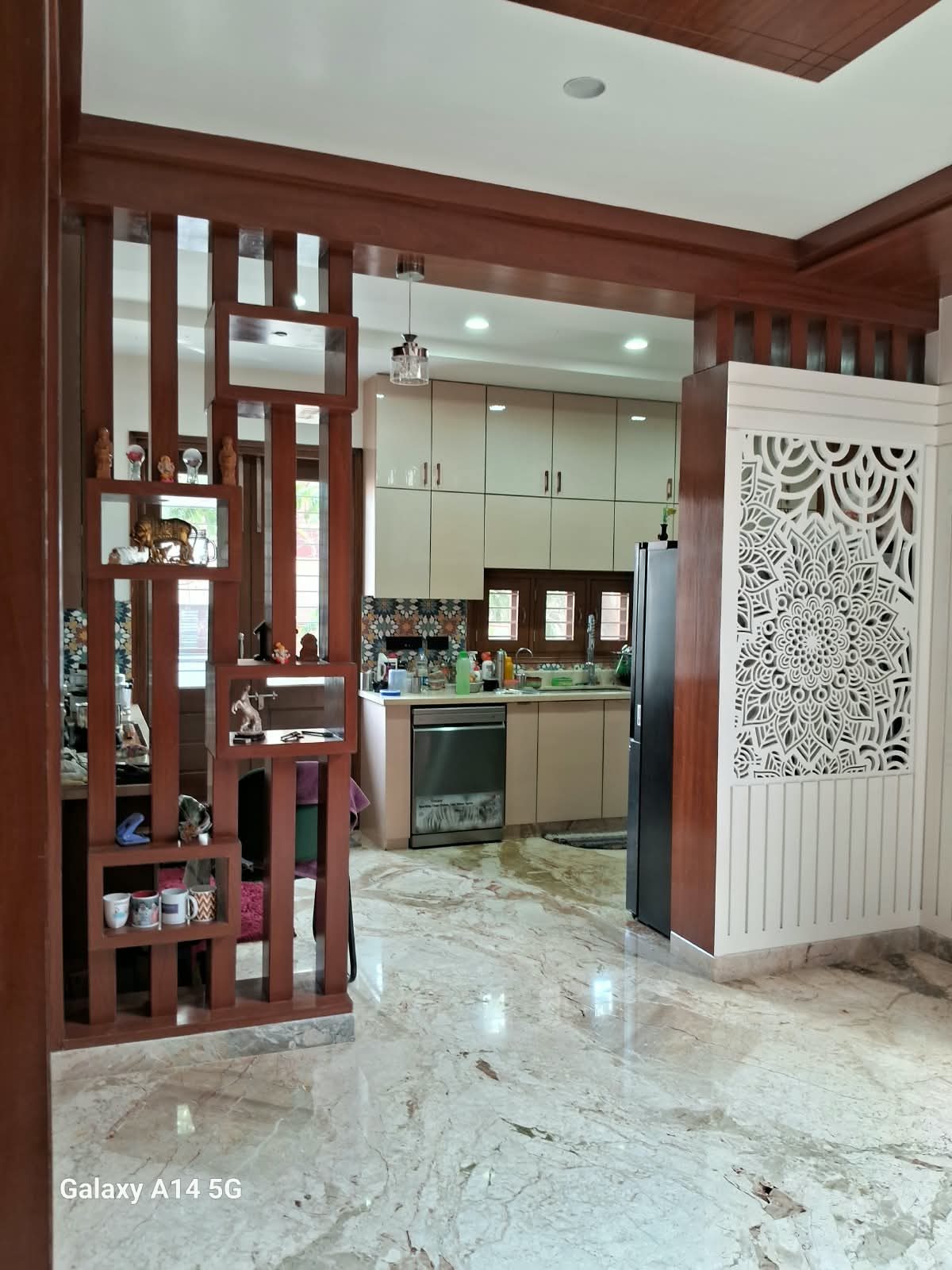 Wood-finish room divider with display shelves beside a carved white jaali panel, kitchen cabinetry behind
